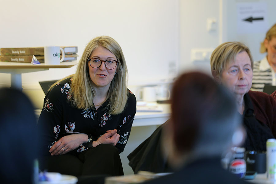 Person med glasögon och blont hår sitter vid ett mötesbord och pratar med en grupp under ett möte i ett kontorsrum.