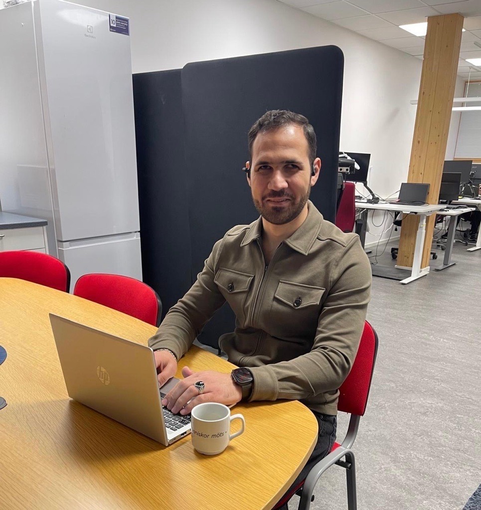 person som sitter vid ett bord framför datorn. på bordet står en laptop och en kaffemugg.