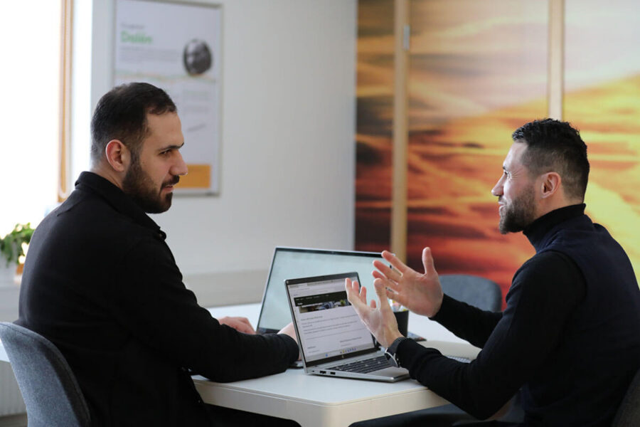 Two people are sitting at a table discussing in front of a laptop in an office environment.