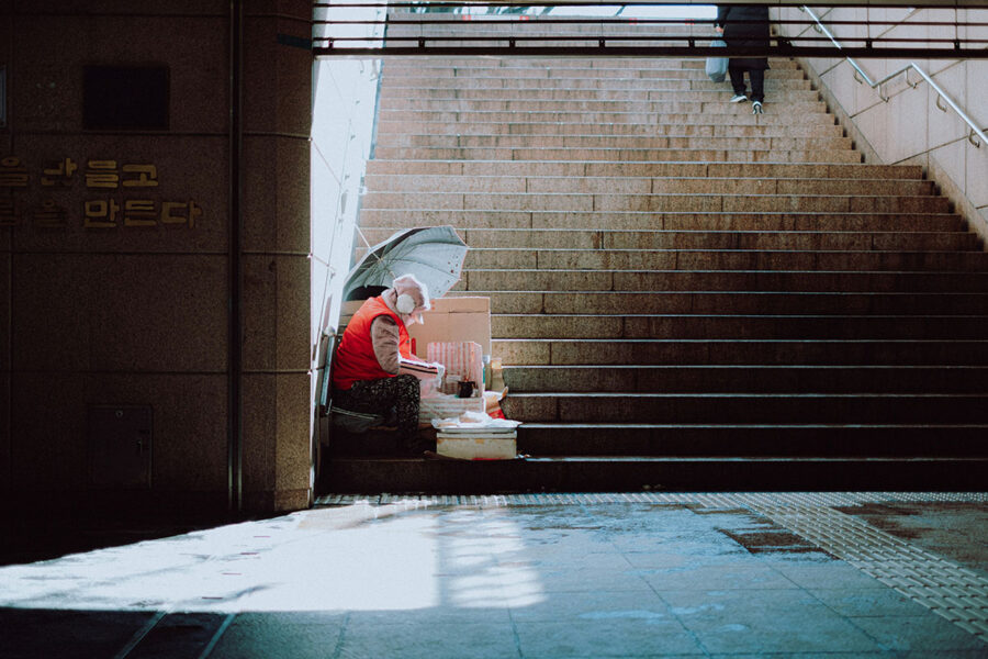 Person in red sweater sitting on stairs in a tunnel holding an open umbrella while bright sunlight shines in from the side.